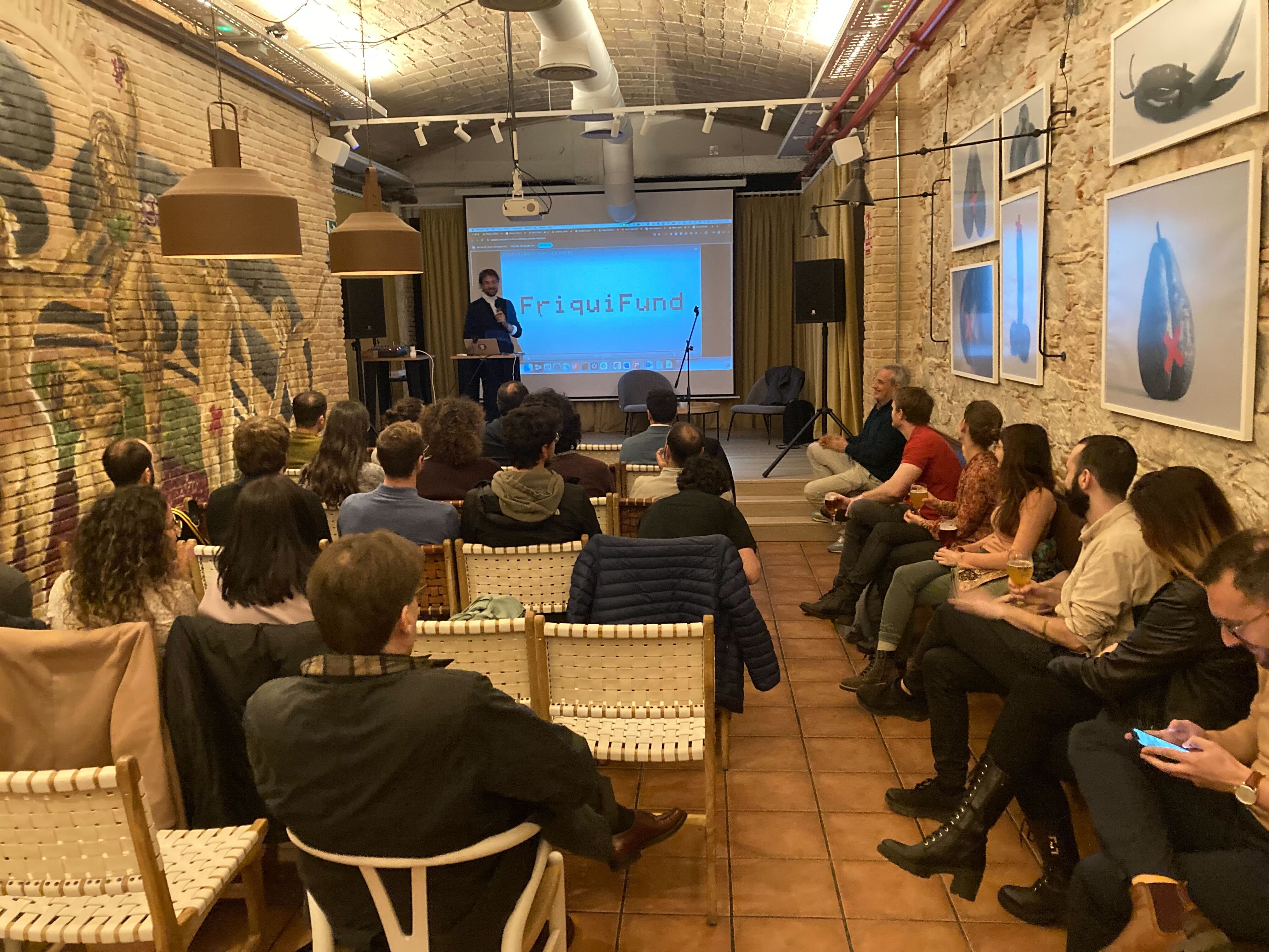 A man standing in front of a screen on which is projected 'FriquiFund' with a small audience seated in front hof him in a hip looking space with art on the wall.